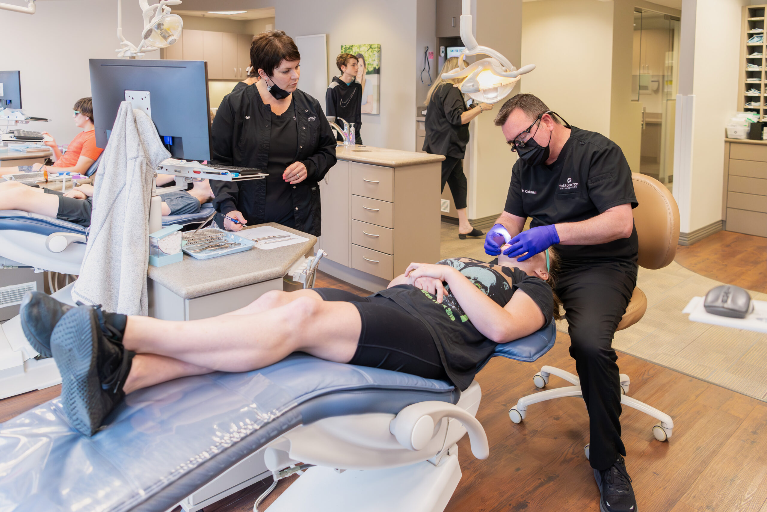 Male orthodontist performs braces treatment on a teen patient in the office while an assistant watches.